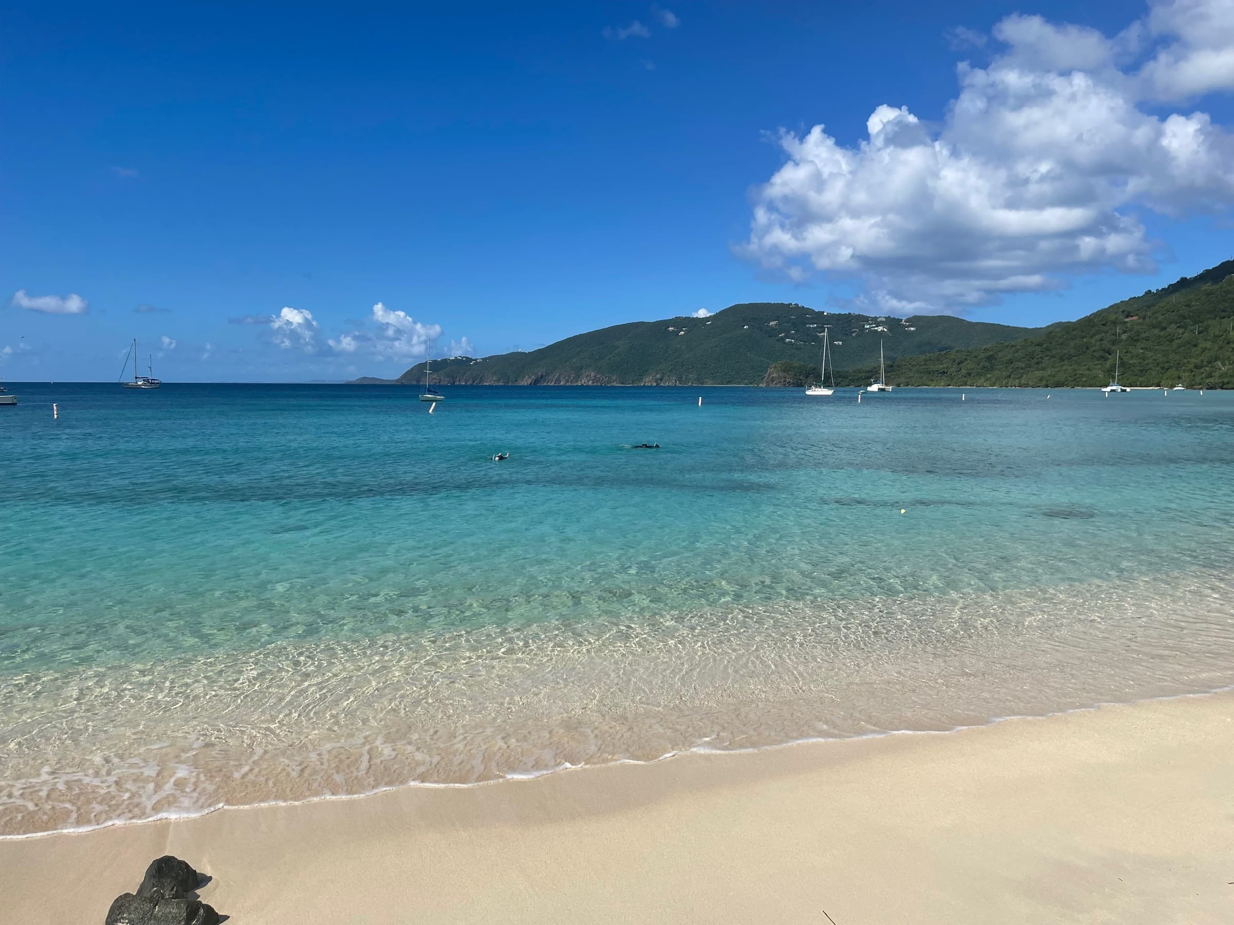 Calm turquoise cove with a white-sand beach, sailboats at anchor, and green hills